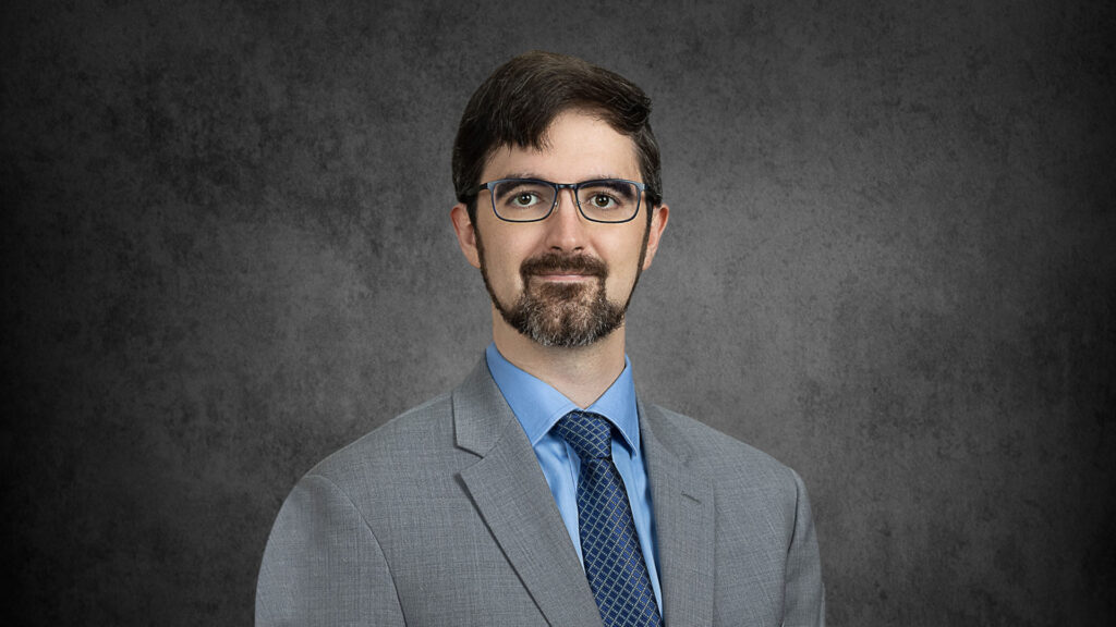 Man with glasses with a beard and moustache wearing a gray suit and blue shirt and tie, in front of a gray backdrop.